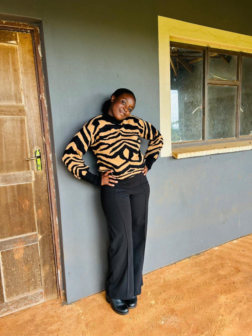 Smiling woman wearing a tiger-print sweater and black trousers posing against a grey wall.