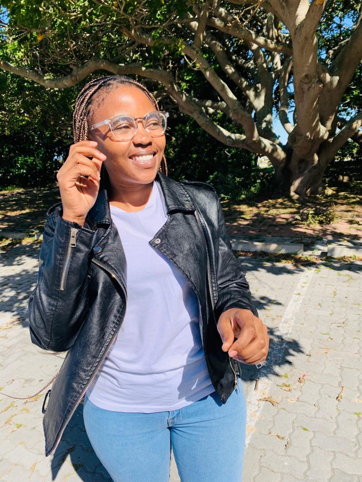 Smiling woman with braided hair wearing a black leather jacket and clear glasses outdoors.