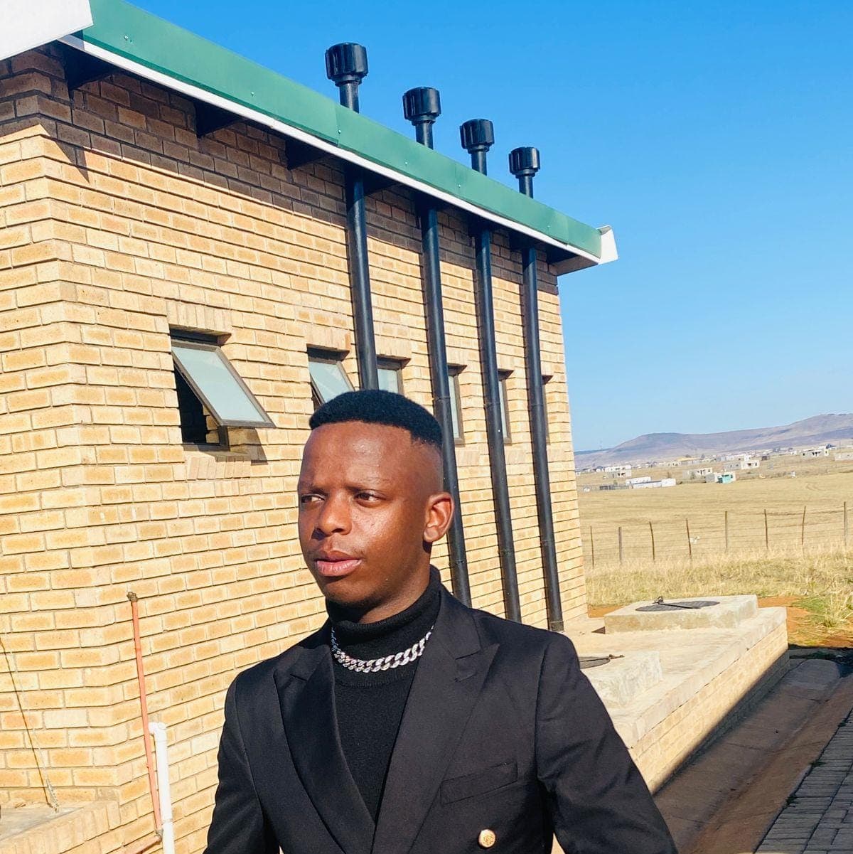 Man in black suit and turtleneck standing outside a brick building under a blue sky.