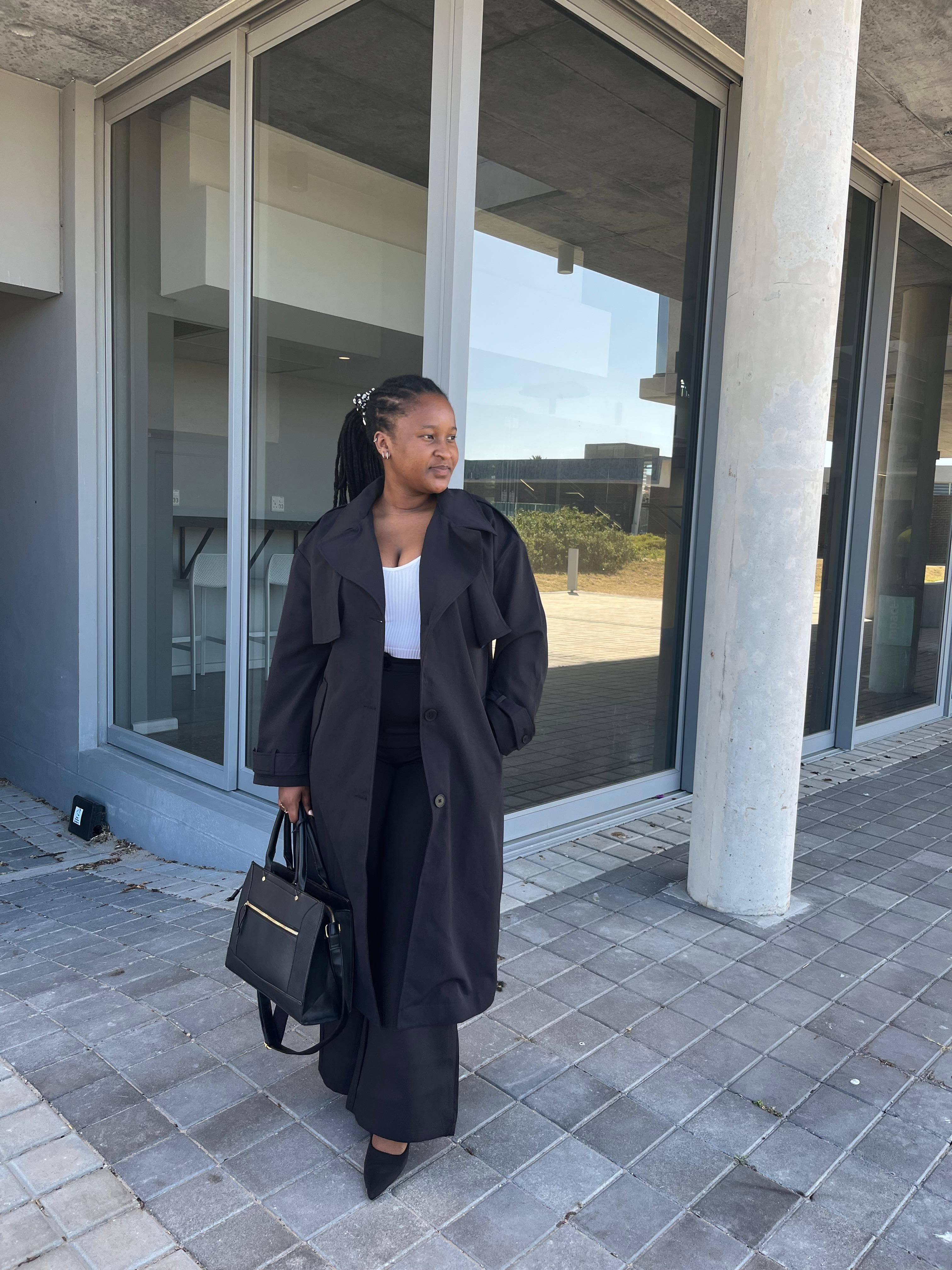 Woman with braids wearing a black trench coat stands outside a modern glass building.