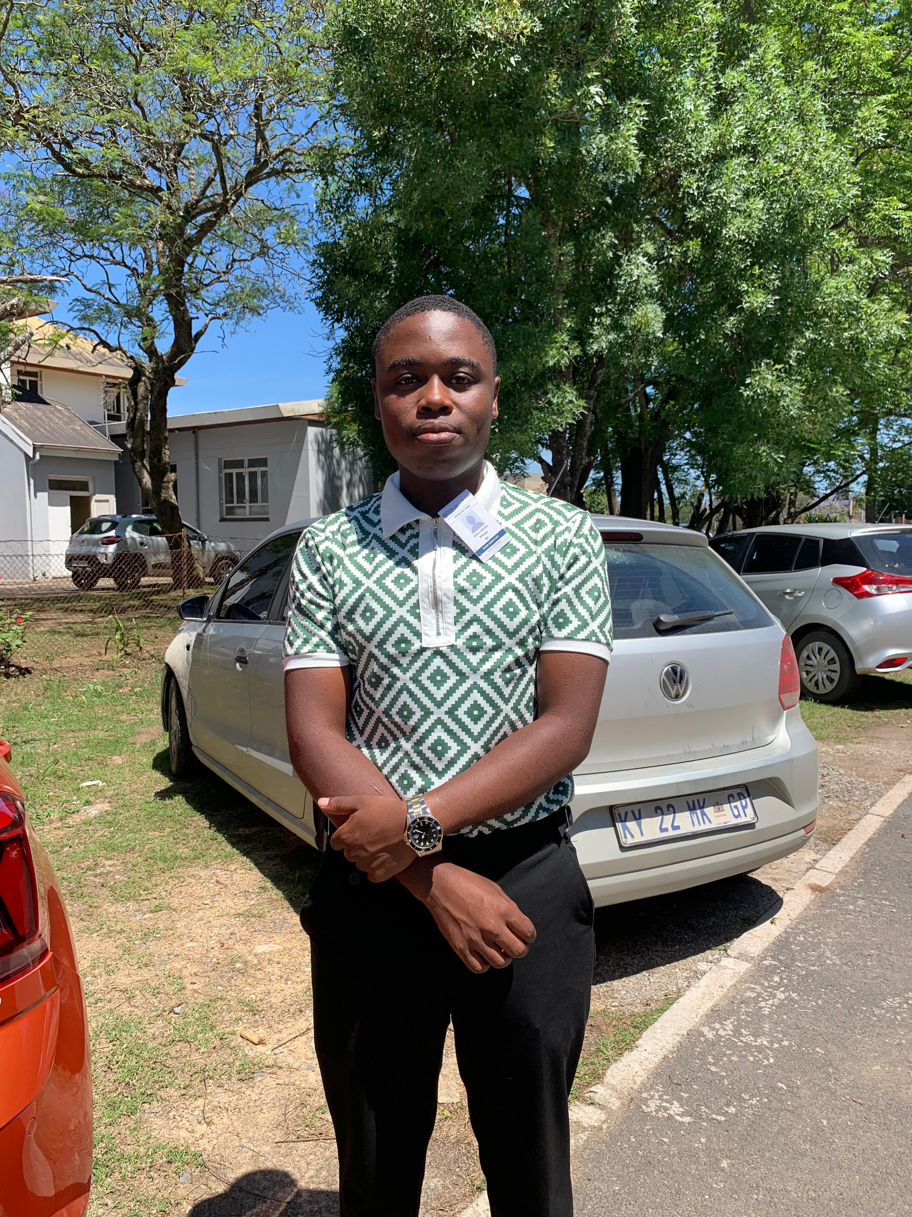 Man in a green geometric polo shirt stands with arms crossed before a silver Volkswagen car.