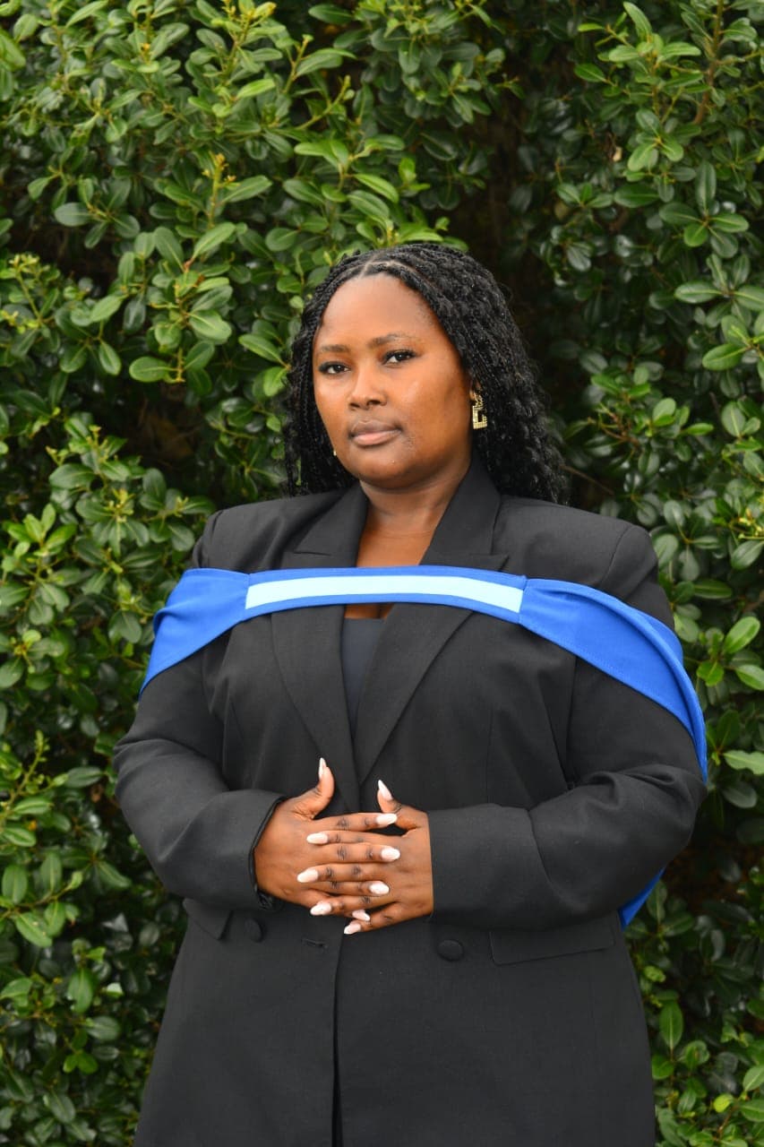 Black woman with braids wearing a black suit and blue graduation hood against green foliage.