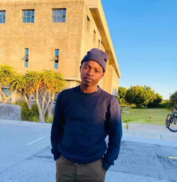 Young man in a blue beanie and sweatshirt stands outside a brick building at sunset.