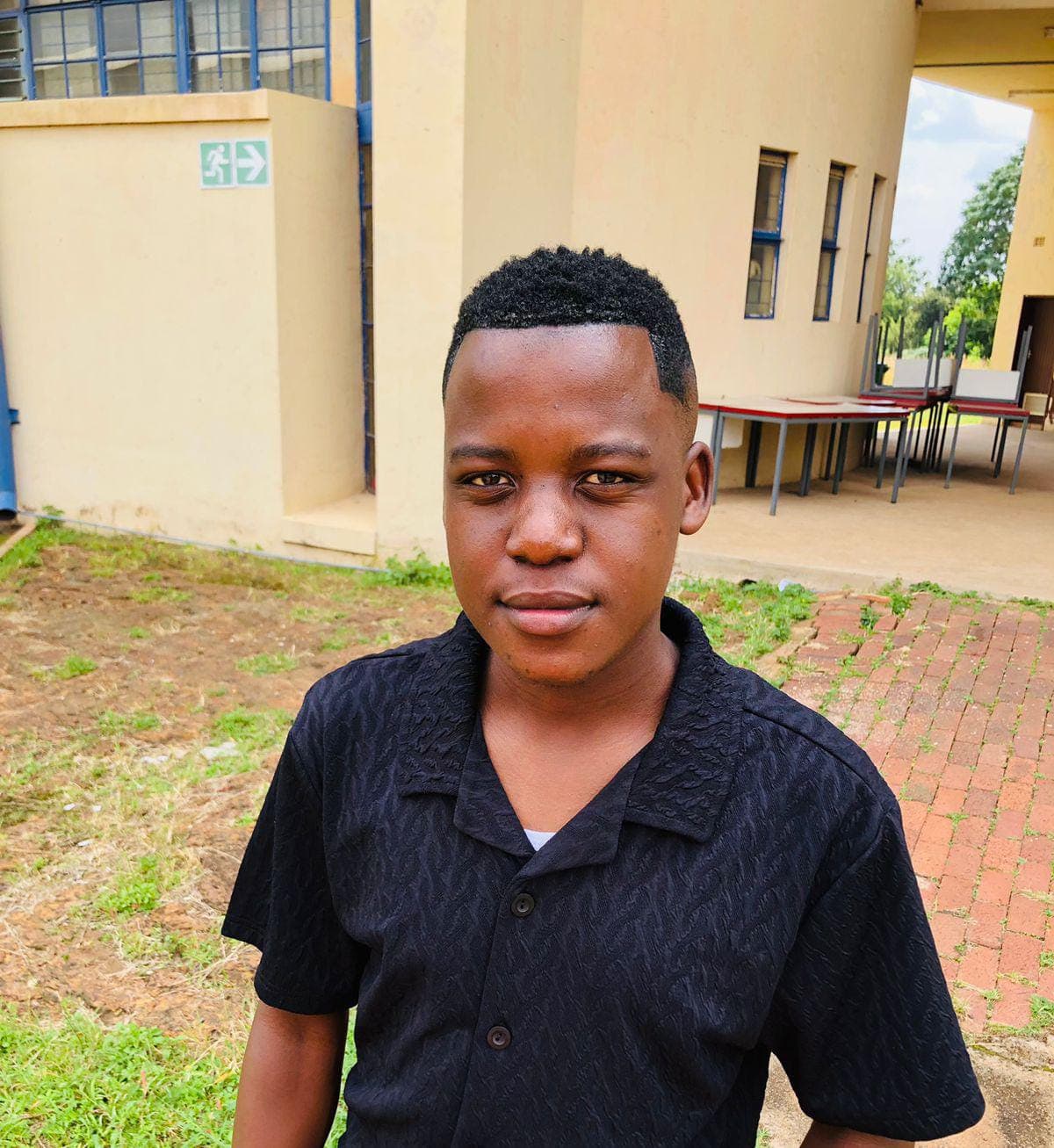 Young Black man with short hair wearing a black textured shirt, standing outside a building.