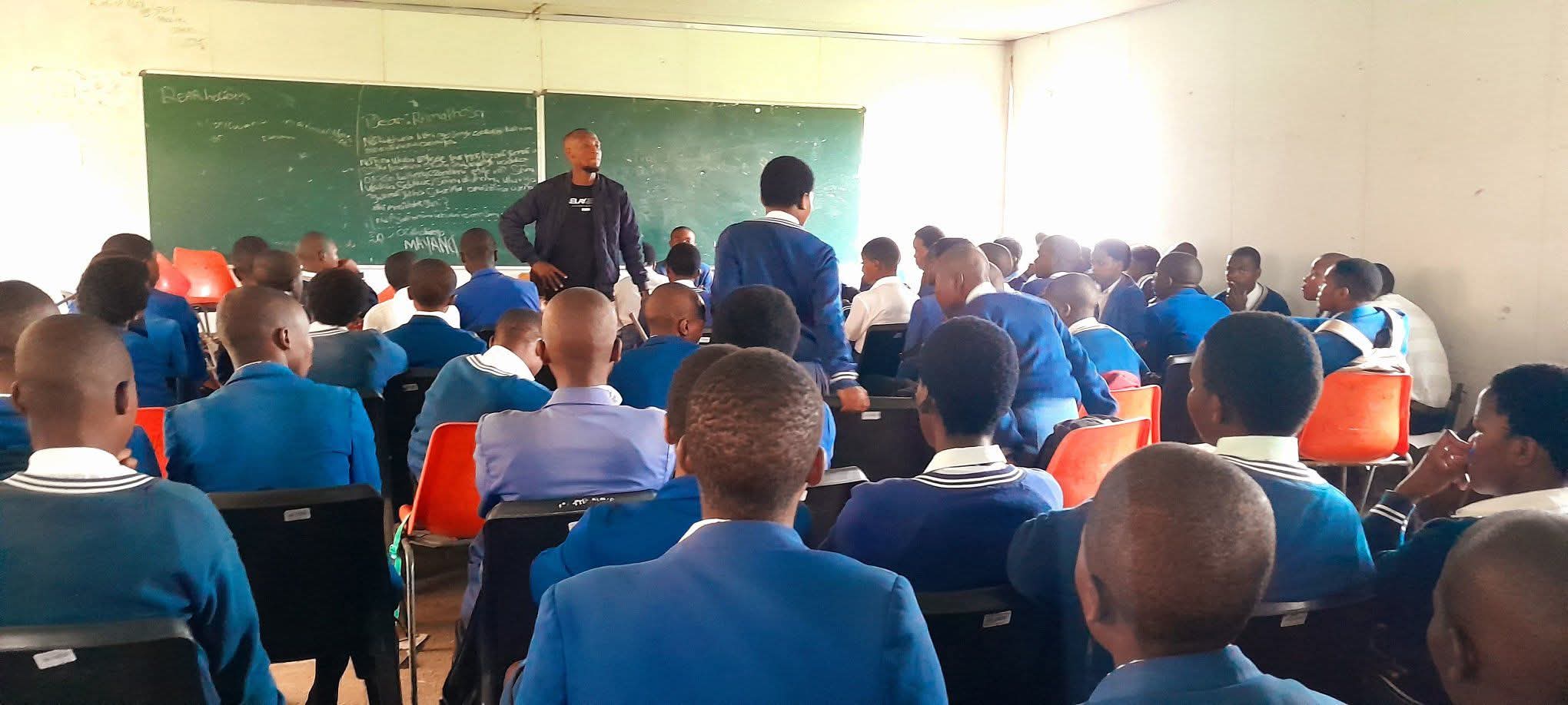 A teacher addresses a classroom of students wearing blue uniforms in front of a chalkboard.