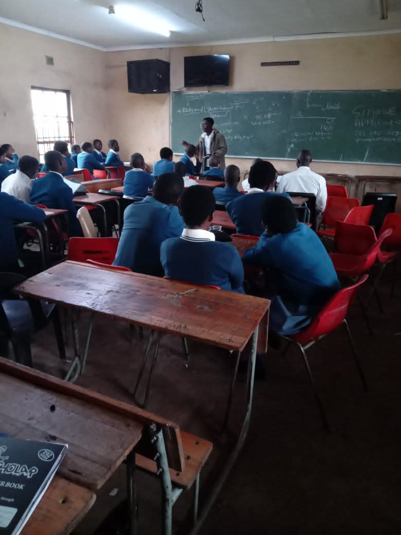 Students in blue uniforms sit at wooden desks facing a teacher at a chalkboard.