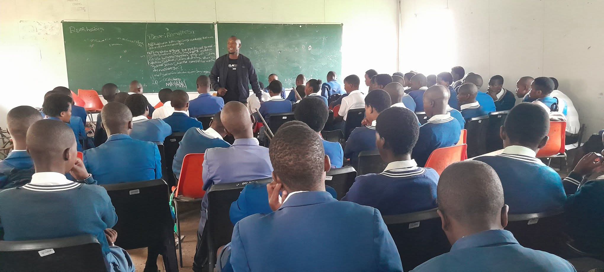A male speaker addresses a classroom of students in blue uniforms before a chalkboard.