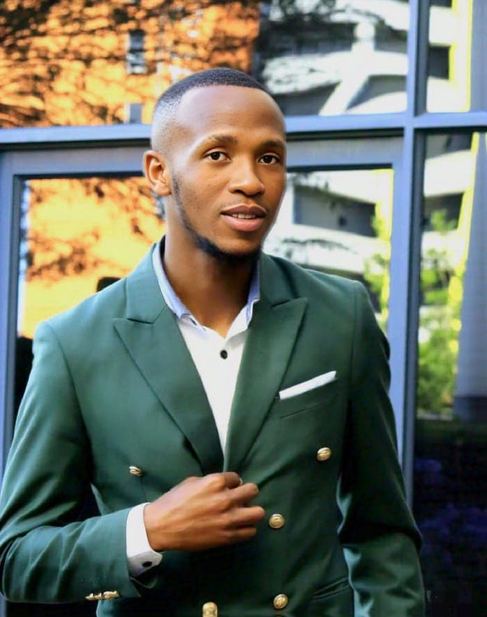 Young Black man in a dark green double-breasted suit with gold buttons posing outdoors.