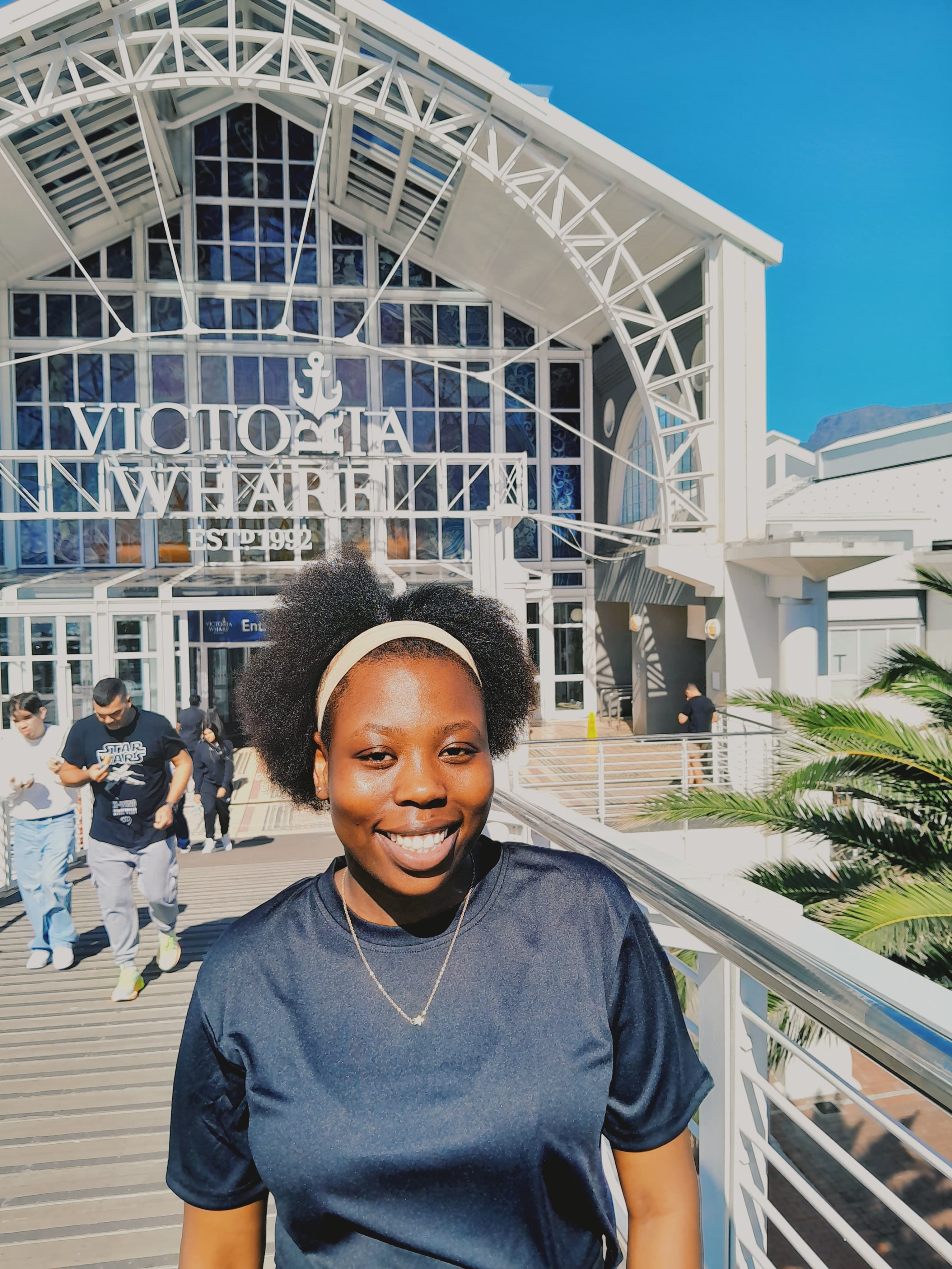 Smiling woman with an afro and headband stands outside the Victoria Wharf building in sunlight.
