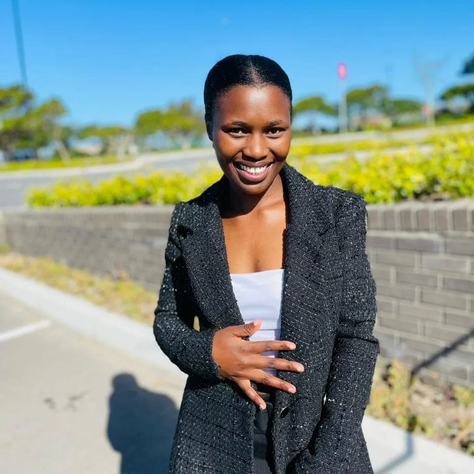 Smiling Black woman in a black textured blazer and white top posing outdoors in bright sunlight.