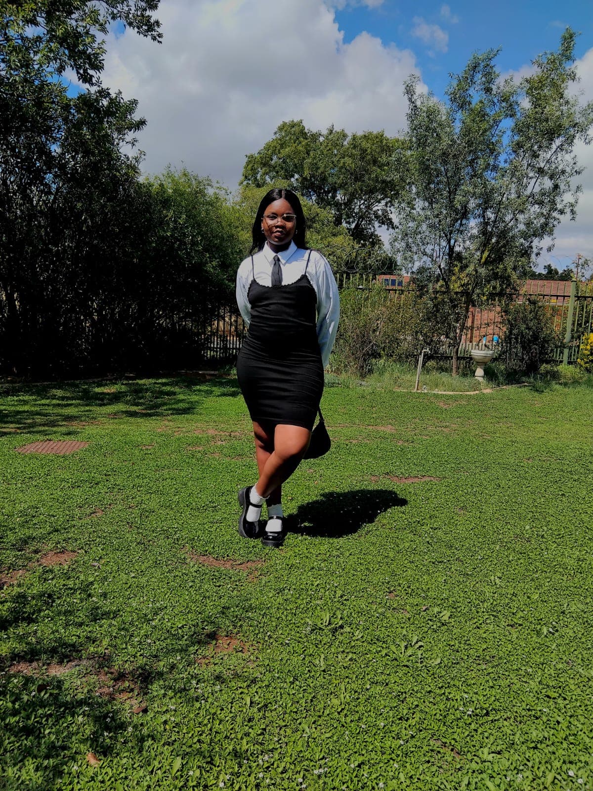 Young woman in a black dress, white shirt, and tie posing on a sunny lawn.