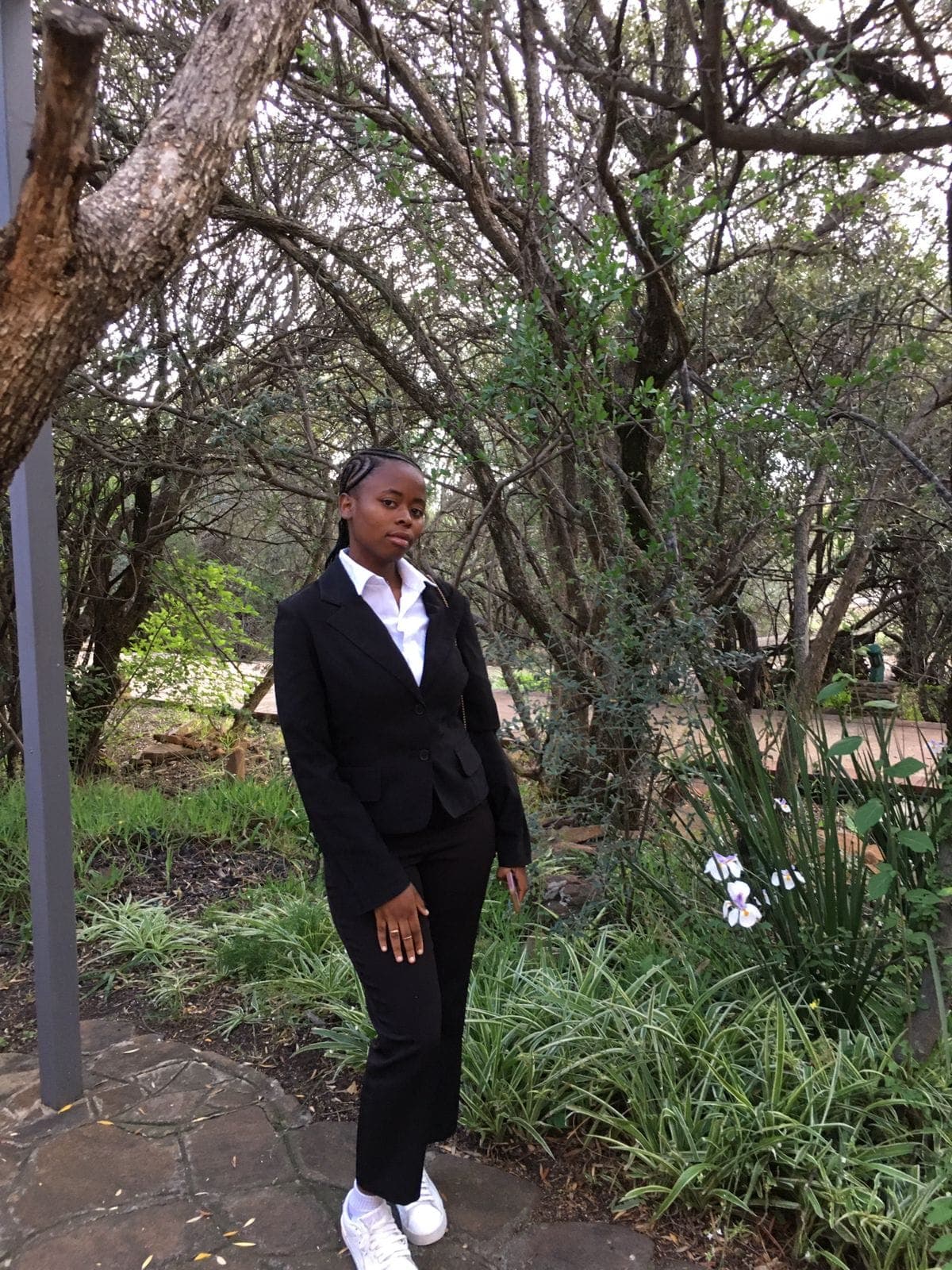 Black woman wearing a professional suit and white sneakers posing in a dense wooded garden.