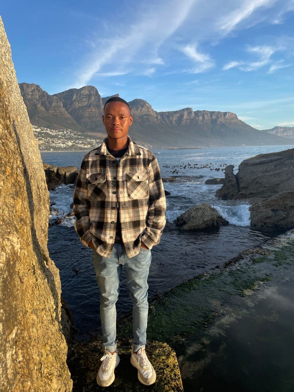 Man in a plaid shirt stands on a rocky shore with mountains in the background.