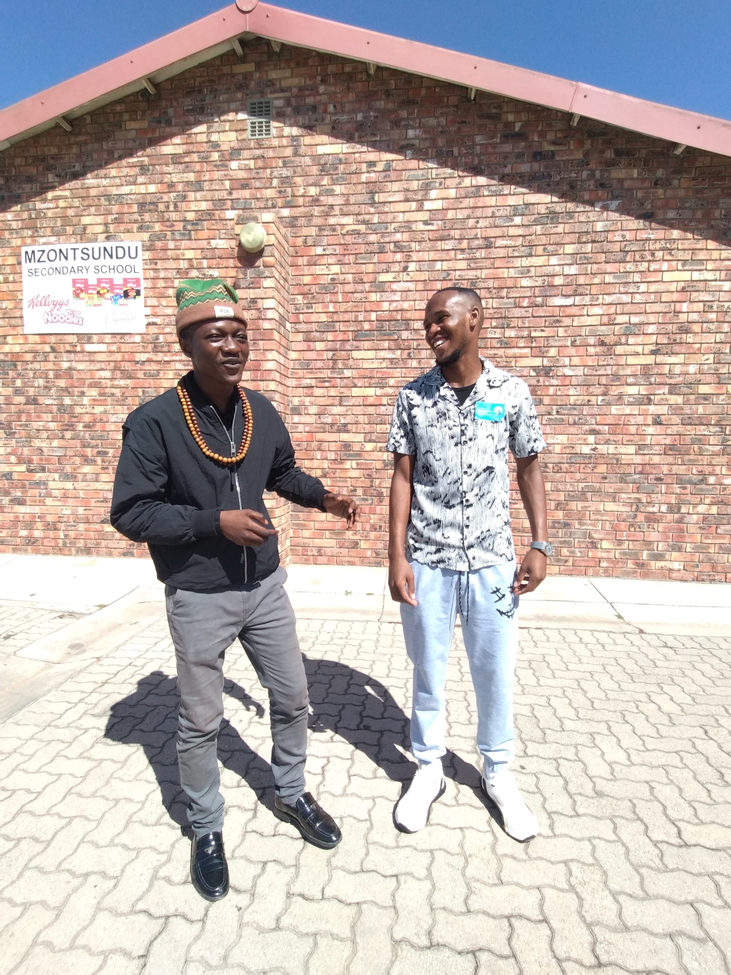Two men talking and laughing in front of Mzontsundu Secondary School's brick wall.