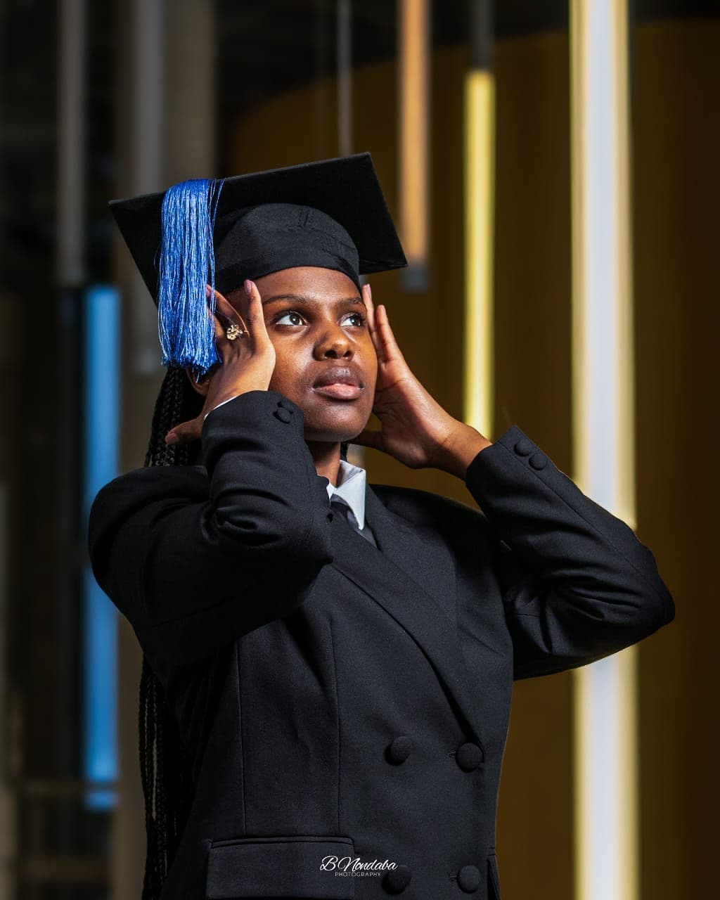 Young Black woman in a graduation cap and black blazer looking upward with thoughtful expression.