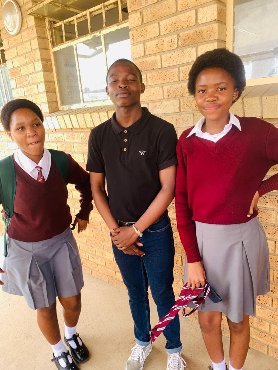 Two girls in maroon school uniforms and a boy in a black polo pose outdoors.