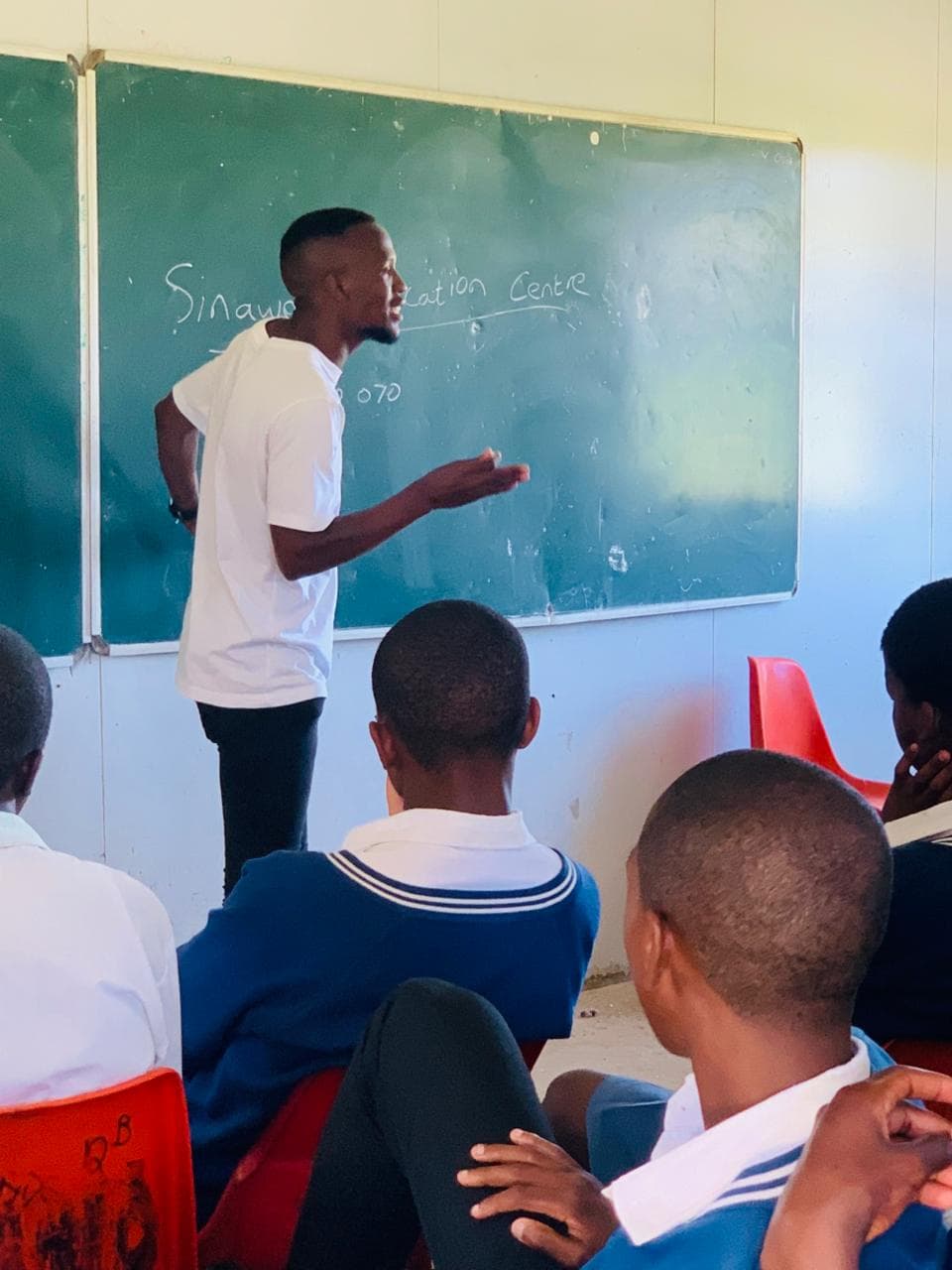 A young man gestures while teaching students in a classroom in front of a chalkboard.