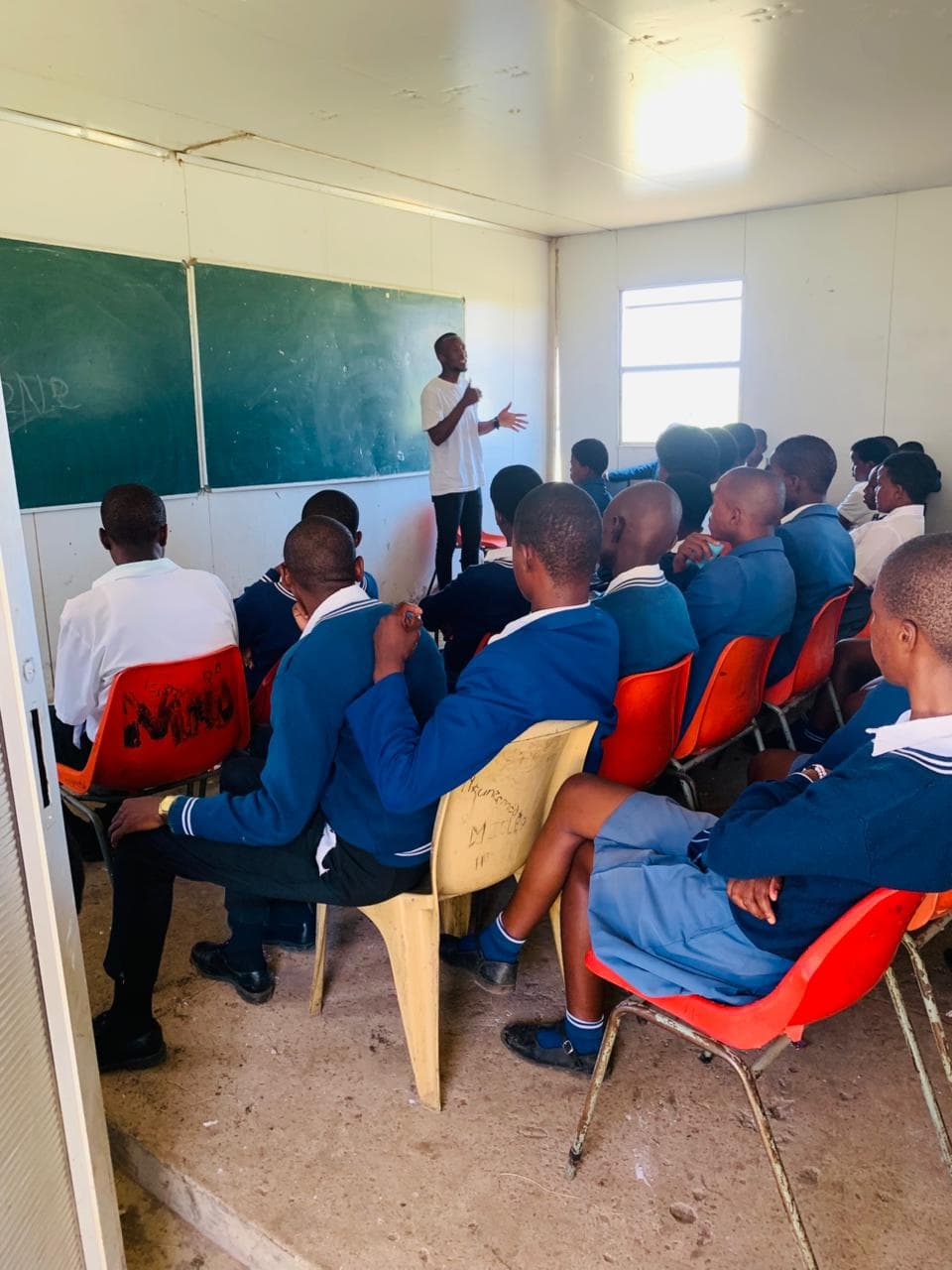 A man addresses a classroom of students in blue uniforms seated before a green chalkboard.